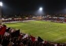 Torcida com bandeiras vermelhas, pretas e brancas em estádio pequeno à noite